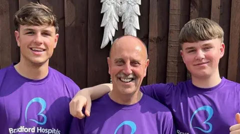 Cara Haigh An older man and two teenage boys stand against a dark fence. They are all wearing purple T shirts with blue logos on.