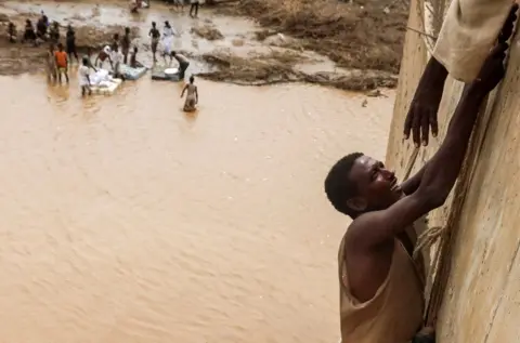 EL TAYEB SIDDIG / REUTERS A survivor is being helped to climb a wall. Behind him is muddy, brown water.