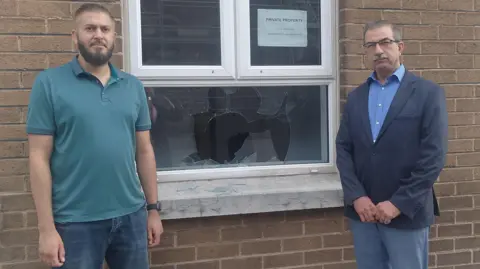 Kashif Akram and Raied Al-Wazzan standing beside the window that was brokem in the attack on the Belfast Islamic Centre. Mr Akram has short brown hair and a beard and is wearing a green polo shirt and blue jeans. Mr Al-Wazzan has short grey hair and a grey moustache. He is wearing glasses, a blue shirt, a navy suit jacket and grey trousers.