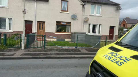 A police van outside a terraced house in Craigton Avenue, Merkinch, in Inverness, where two people were seriously injured. There is a lamp post with a 20mph speed limit sign on it, and wire fences along the front of gardens. 