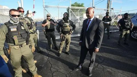 Mayor Baraka, seen visiting the same facility on Wednesday. He is a black man, and wears a dark suit. Armed officers wearing masks gather around him