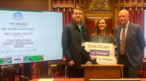 Ciarán Mac Giolla Bhéin with two other people at an event for Irish language week. The woman in the middle is holding up a sign saying 'Seachtain na Gaeilge le energia'.