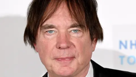 A man with dark hair that has been brushed to either side of his face is pictured looking expressionless in front of a white background. He is clean shaven and has greyish blue eyes.
