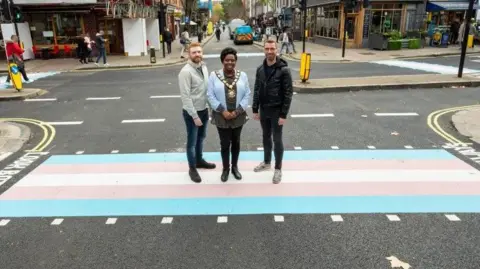 Camden Council Three people stand on a road crossing in a London street: the crossing is painted in blue, pink and white stripes. They are casually dressed men and a woman who is wearing a blue jacket and a mayoral chain of office.