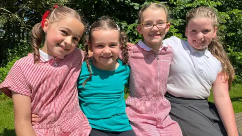 Freya sits next to her sister Gracie and friend Alya and Emily. They are wearing their school uniforms from Hartside Primary Academy. 