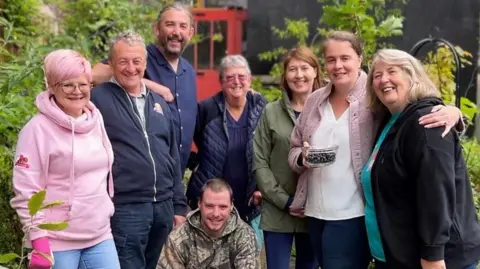 Martin, Paula and community volunteers pictured for BBC Radio Manchester's Make a Difference Awards 2023 : a group of five women and three men stand smiling, with their arms around one another outside; one man kneels at the front of the circle.