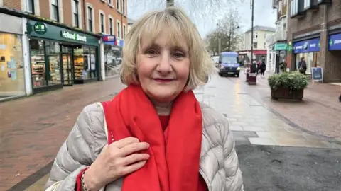 A blonde woman wearing a light grey coat and a red scarf. She is standing in the middle of a high street.