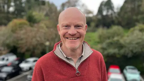 BBC Deputy Gavin St Pier - A man with a bald head and a ginger and grey beard smiling at the camera in a car park. He is wearing a red jumper. 