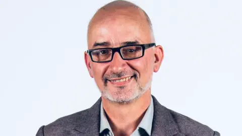 Leslie Laine A man wearing black-framed glasses, he has a black and white beared and is smiling. He is wearing a black suit with a grey shirt.