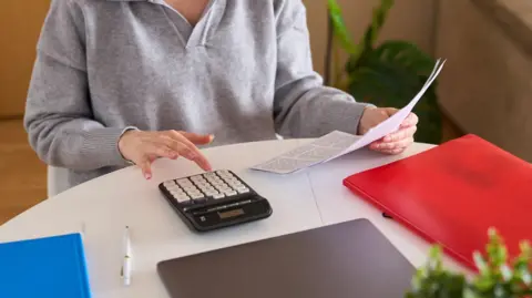 A woman is wearing a grey jumper and glasses. She is sat at a desk and is looking at a document and is using a calculator. 