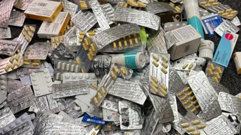 A large pile of pharmaceutical products scattered on a dark carpeted floor.
The central focus is a heap of medication blister packs, many of which contain yellow-and-white capsules.