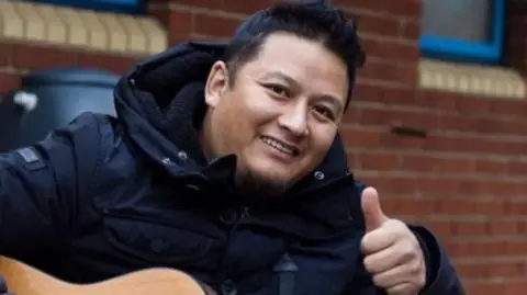 A man with dark hair and a dark winter coat smiles and puts his thumb up while holding a guitar on his knee.