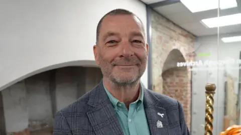 Qays Najm/BBC Simon Ring standing inside a council building. He is looking directly at the camera and smiling. He is wearing a light teal coloured shirt, with a grey blazer over the top.