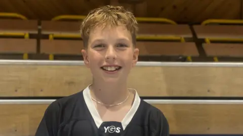 Archie is a young, fair-skinned boy with blonde hair wearing a black jersey with white on the neck. He is standing in the roller rink with a side wall behind him.