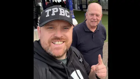 Scott Johnson A man in a black hoodie and black hat, and a man in a black polo shirt are standing in front of Eastbourne Pier.