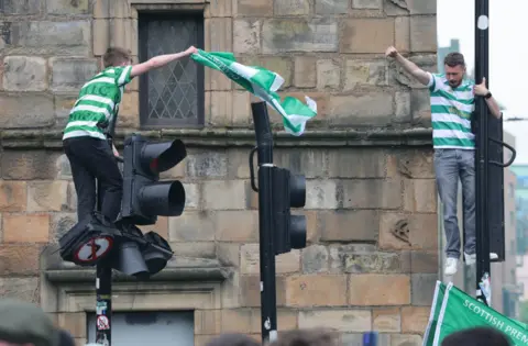 PA Media Two Celtic fans climb traffic lights and wave a green-and-white flag during title celebrations in the city centre.