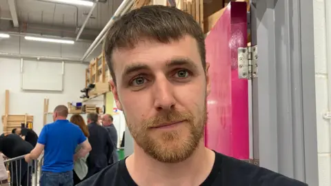 Aaron Emerson has short brown hair and blue eyes, he also has a light brown, blonde beard. He is wearing a black t-shirt and is standing in front of a crowd watching people compete at the competition.