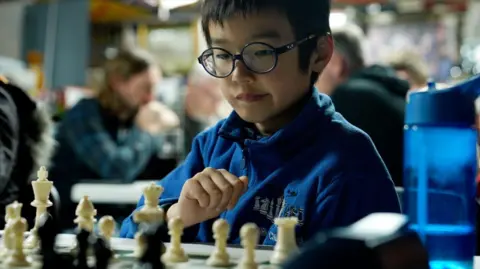 Jayden is wearing a royal blue zip-up fleece and round, black-rimmed glasses. He is concentrating on a chess board and has one hand up, poised, ready to move a piece. Out of focus behind him are other, adult, players sitting at a different table.