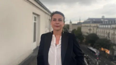 Elodie Tuaillon-Hibon is photographed on a balcony in Paris. She is looking straight into the camera and wearing a white shirt and black jacket with large gold earrings. 