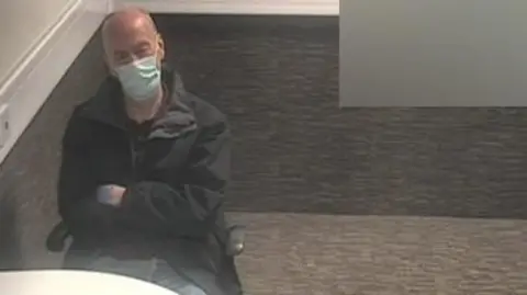 Greater Manchester Police An older man sits with his arms crossed and wearing a face mask in a carpeted room in a police station. 