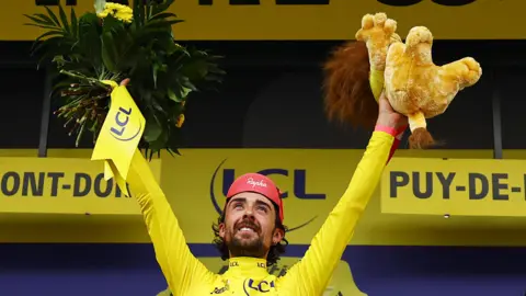 Reuters/Sarah Meyssonnier Ben Healy celebrates on the podium wearing the yellow jersey after stage six of the Tour De France. He is holding a stuffed animal in his left hand and flowers in his right.