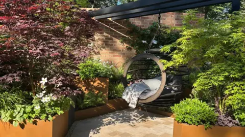 Different planting containers on different levels are full of green plants, some trees with purple leaves and white flowers. In the middle is a hanging chair.