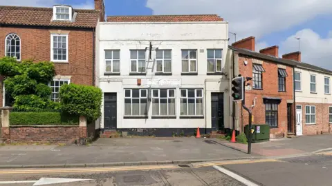 An unused pub. It is a white two-storey building with large windows and two black doorways – one at each end. It is set back from the road with a small forecourt outside.