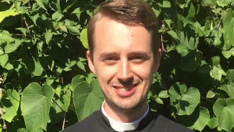 Diocese of Chichester A man with short brown hair and wearing a clerical collar smiles in bright sunlight in front of a green hedge.