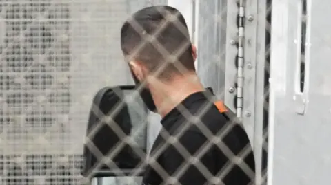 Eddie Mitchell A man in a white uniform is next to a man in dark clothing. They are pictured behind a grey wire fence.