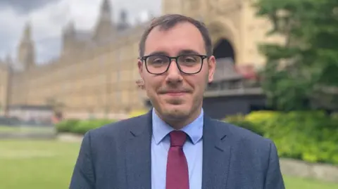 MP Tom Gordon stands outside in front of a large brick building. He wears a suit and tie. 