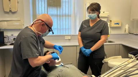 Inside Torrington Dental Practice. A man in a dentist chair in a grey jumper. Over him, to the left, is a dentist wearing grey scrubs and blue gloves and goggles and a blue mask. To the right is a woman with her brown hair tied back, wearing blue gloves and grey scrubs, a mask and goggles. 