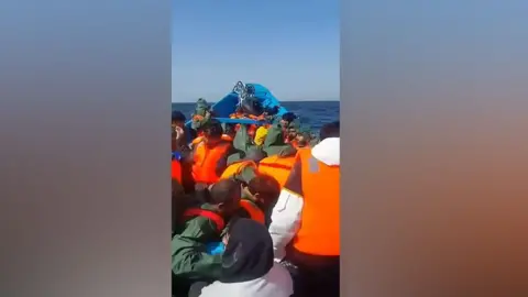 A screen of the boat shortly after leaving Mauritania. There are many immigrants who wear orange survival jackets, all face the camera.