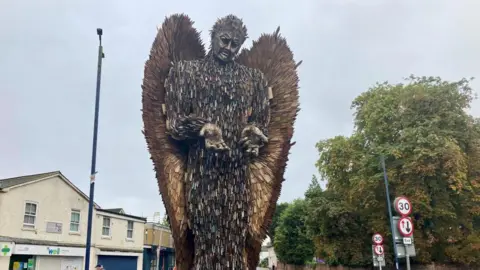 Luke Deal/BBC The Knife Angel statue rises into the sky. It is composed of thousands of knives. In the background are trees and a row of shops. 