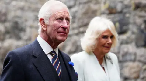 Getty Images King Charles and Queen Camilla walking with a stone wall in the background. He is wearing a suit with a blue striped tie. She is wearing a white dress.