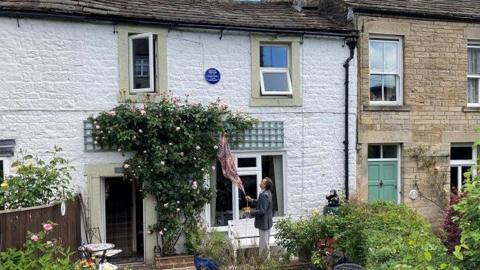 Man in front of a cottage unveiling a blue plaque to mountaineer Bentley Beetham