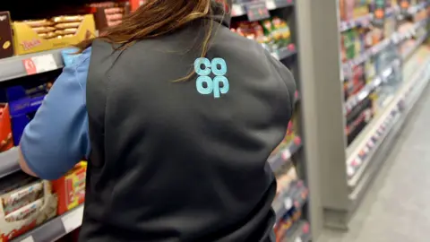A woman with a long brown ponytail wears a black gillet with a blue co-op logo while looking at a shelf full of chocolates in the supermarket.