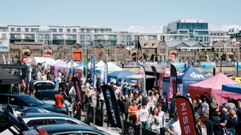Ports of Jersey High angle photo of a bustling pier at Jersey Boat Show. Lots of people are walking up and down a sunny promenade as pop up business tents and flags line the periphery