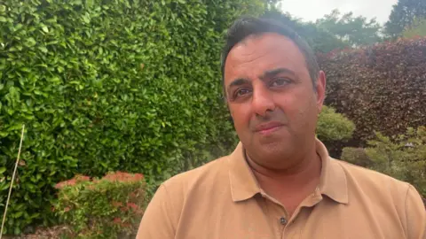 A close-up of a man standing in front of a large hedge. He wears a light brown shirt.