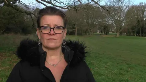 Tina Quadrino looking at the camera with black framed glasses on her face, her hair pulled back and a black coat on with a fur collar. Greenery from the golf course in the background.