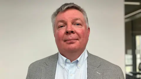 Shariqua Ahmed/BBC Head and shoulders image of Andrew O'Neil. He has short grey hair and is wearing a black and white suit with light blue chequered shirt.