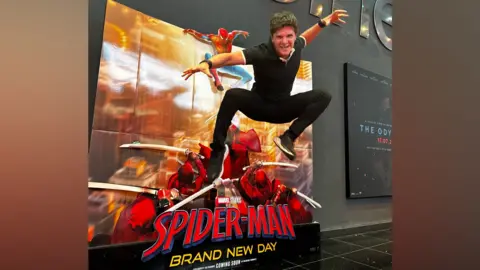 Teifion Sussex A man jumps up and strikes a pose with his arms outstretched. He is in front of a sign reading 'Spider-Man - Brand New Day' against a grey cinema lobby wall
