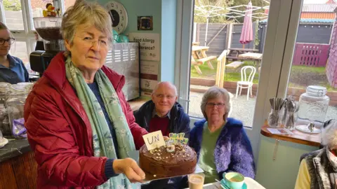 Heather Kidd has short grey hair and is wearing glasses, a red coat and green scarf. She is standing in a cafe and is holding a chocolate cake. The cake has a candle and writing on it that reads unhappy 5th birthday. There is a man and a woman sitting at a table behind Ms Kidd.