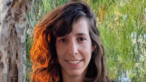 Noa Dagoni A woman with brown hair smiles at the camera with a tree beind