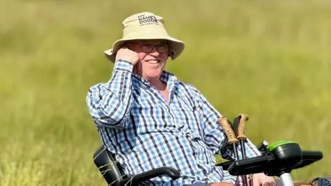 Supplied Nick Apperley sitting on a mobility scooter in a field. He is wearing a hat and a long-sleeved shirt and glasses. He is smiling but looking away from the camera. 