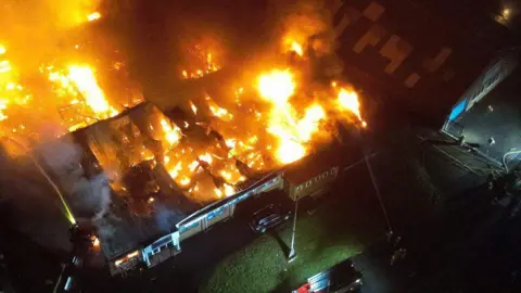 An aerial photo of a blaze at a strip of commercial buildings - the roof has been mostly burnt away showing fierce flames underneath.