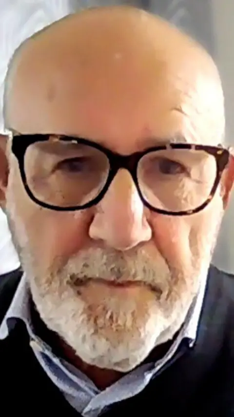 Jon Trickett is in a blue shirt, black jumper and black glasses. He is a bald man with a grey goatee
