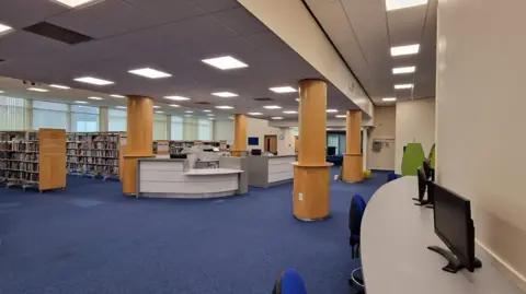 Large open-plan public library with blue carpet flooring, rows of book shelves, curved white service counters, and tall wooden columns supporting a high ceiling with bright panel lighting. Computer workstations line the right side of the space, and large windows along the left wall provide natural light.