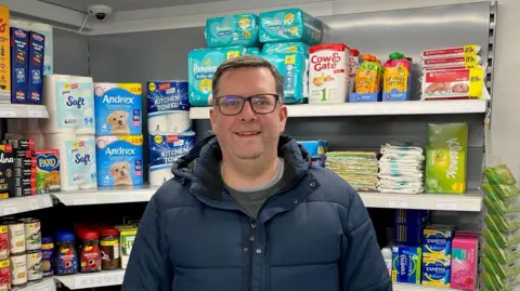 JOHN ATTRILL John Attrill has short brown hair and has glasses on. He is wearing a blue puffer jacket over a grey sweater and light grey shirt. Behind him are items on the shelves in a shop.
