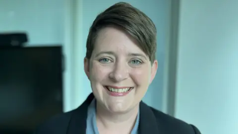 Headshot of education minister Olivia Bailey. She wears her cropped brown hair parted and swept to one side. She is wearing a powder-blue shirt and a black jacket. 