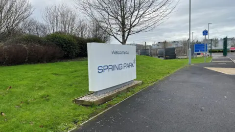 A prominent concrete sign outside a former MOD base with the words "Welcome to Spring Park" written on it. Fencing and further signs can be seen in background.